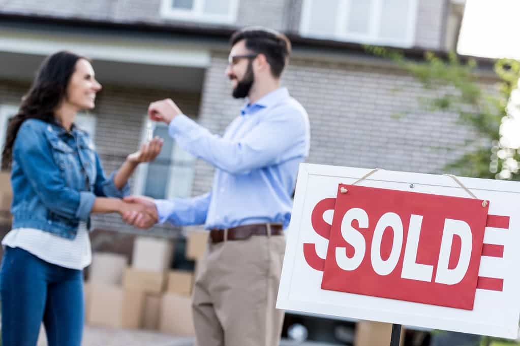 Realtor in front of a large home handing a woman keys behind a Sold sign
